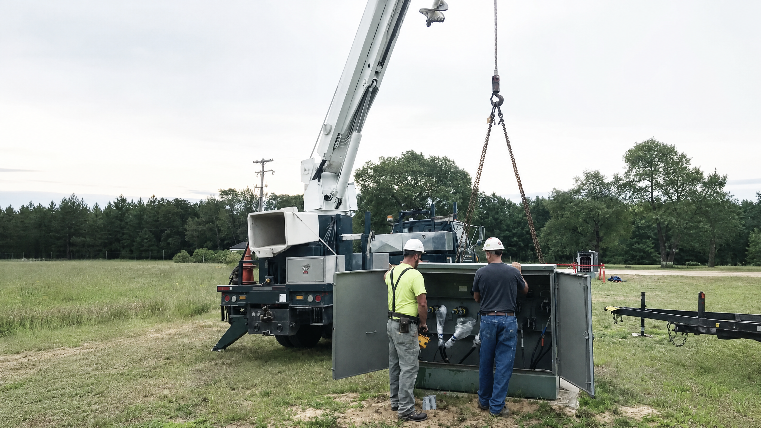 Two workers beside an open padmount transformer and an unbranded crane truck