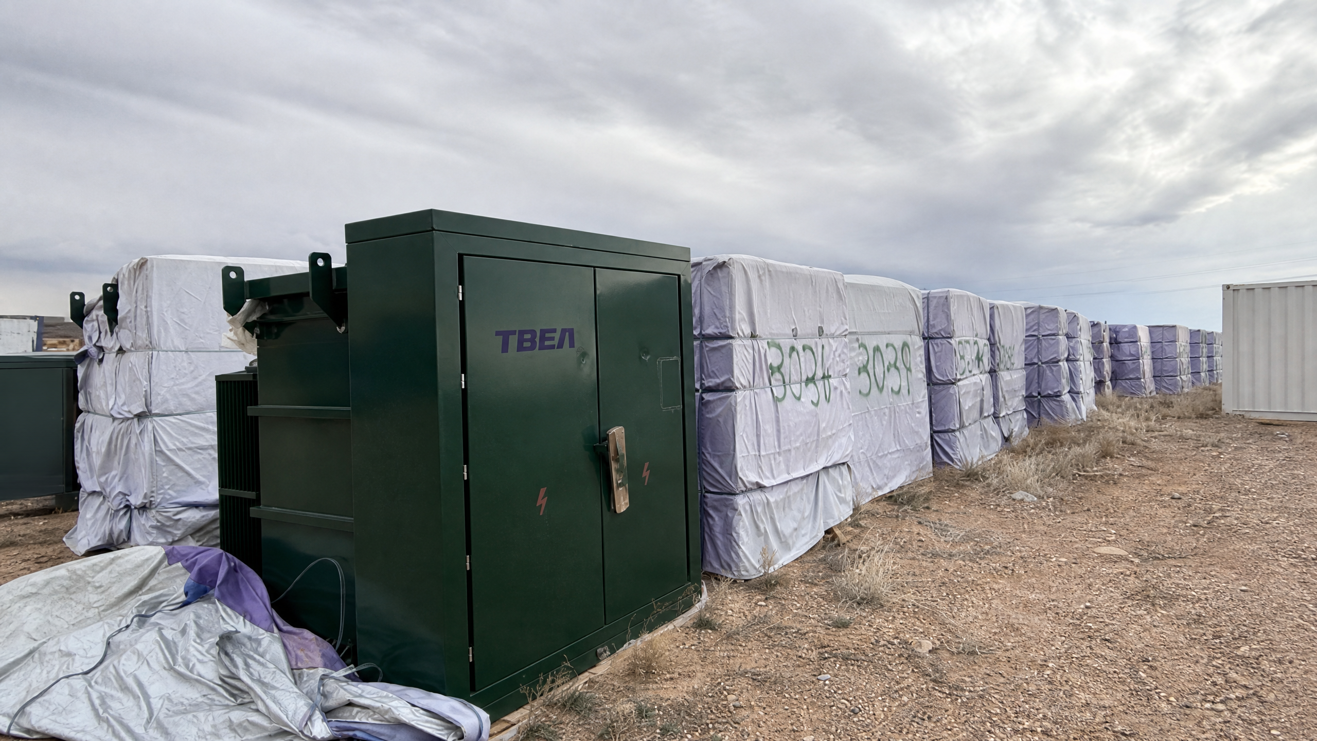 Dark green padmount transformer beside wrapped equipment in an outdoor field