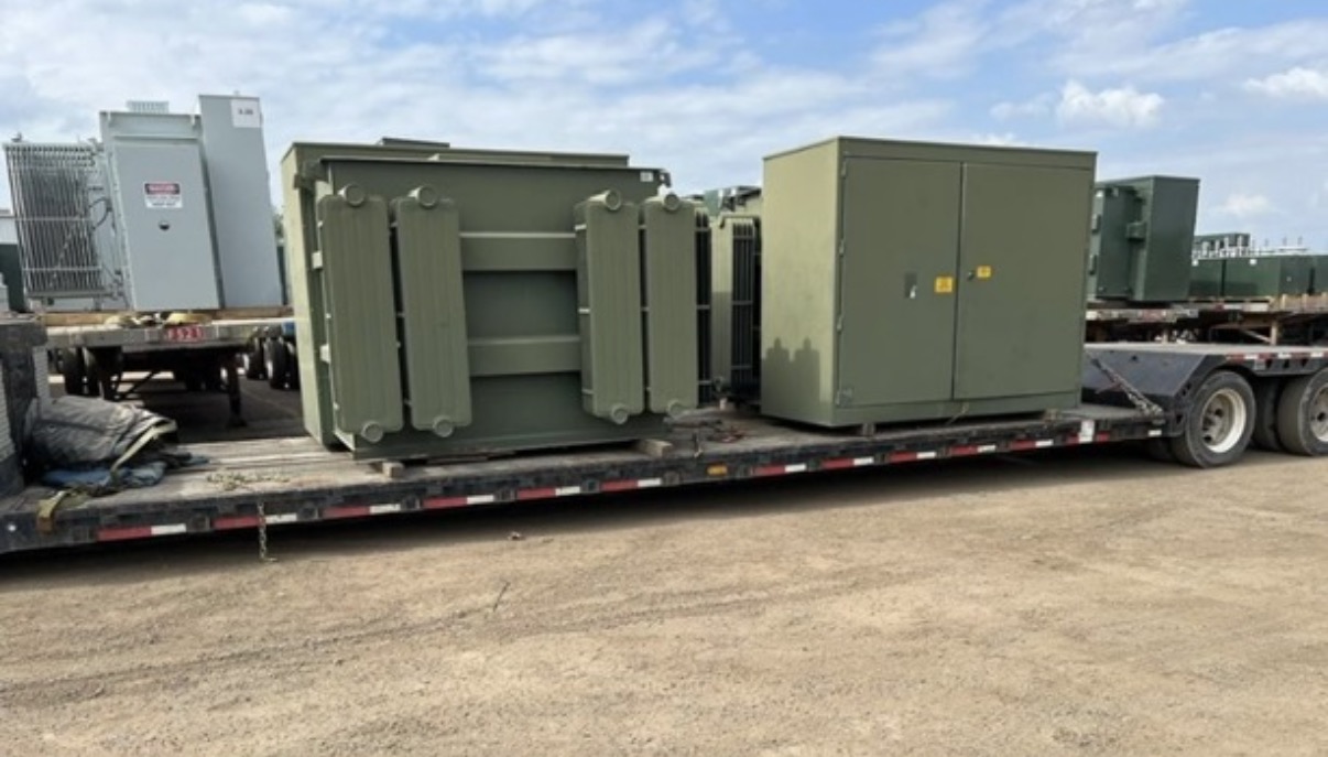 Large green padmount transformers secured on a flatbed trailer in an outdoor yard