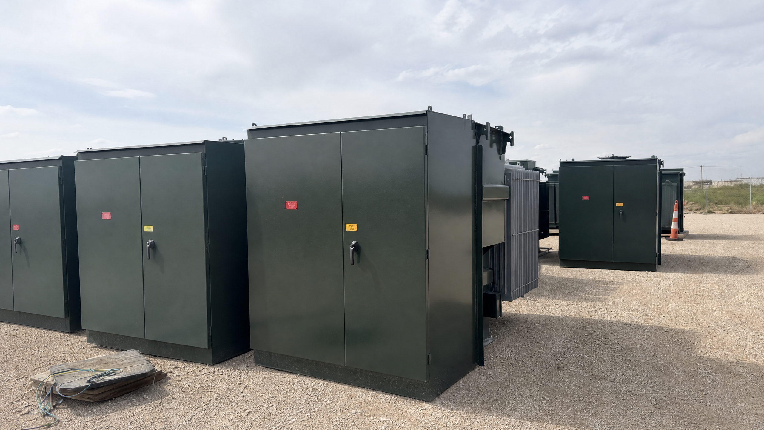 Multiple green padmount transformers lined up in an outdoor gravel yard